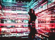 Image of a man kneeling inside a building, with neon lights and reflections, to suggest many dimensions of AI. Image by Drew Dizzy Graham, Unsplash, https://unsplash.com/license